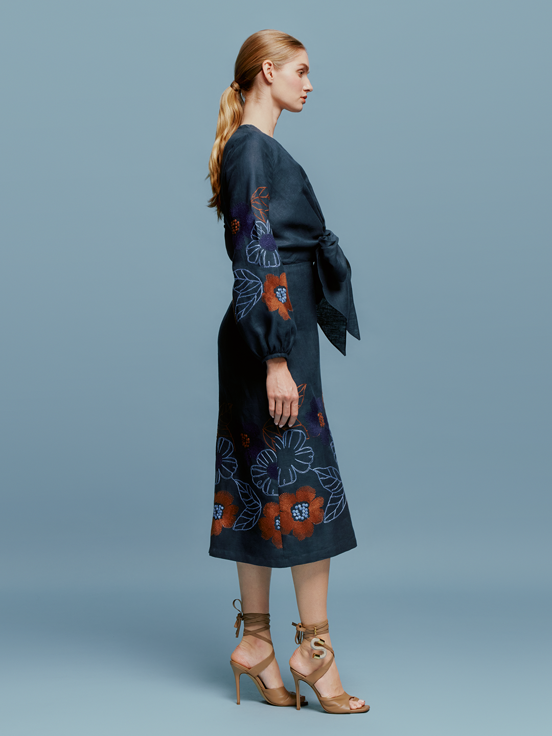 A person stands against a blue background wearing the Laoise Dress in Navy Ochre Flower Embroidery, paired with beige high-heeled sandals, perfect for an August 19th or September 2nd outing.