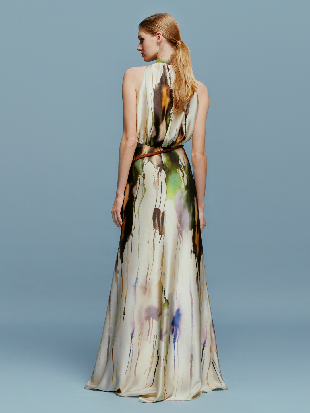 A woman in an elegant, long, Mabel Dress Olive Watercolor with an abstract design, available for pre-order, standing against a light blue background.