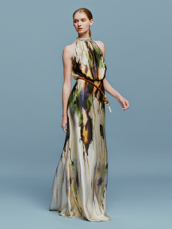 A woman in an elegant, long, Mabel Dress Olive Watercolor with an abstract design, available for pre-order, standing against a light blue background.