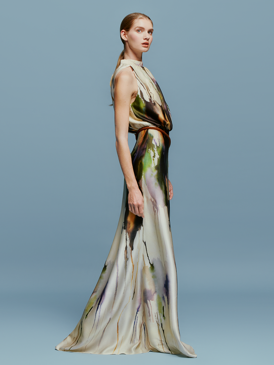 A woman in an elegant, long, Mabel Dress Olive Watercolor with an abstract design, available for pre-order, standing against a light blue background.