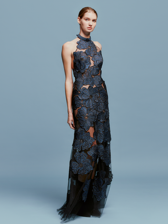 A woman is elegantly adorned in the Mab Dress Navy, featuring a halter-neck style with large floral blue appliques and sheer fabric, standing against a plain blue background, capturing the sophisticated essence of an August 19th gala.