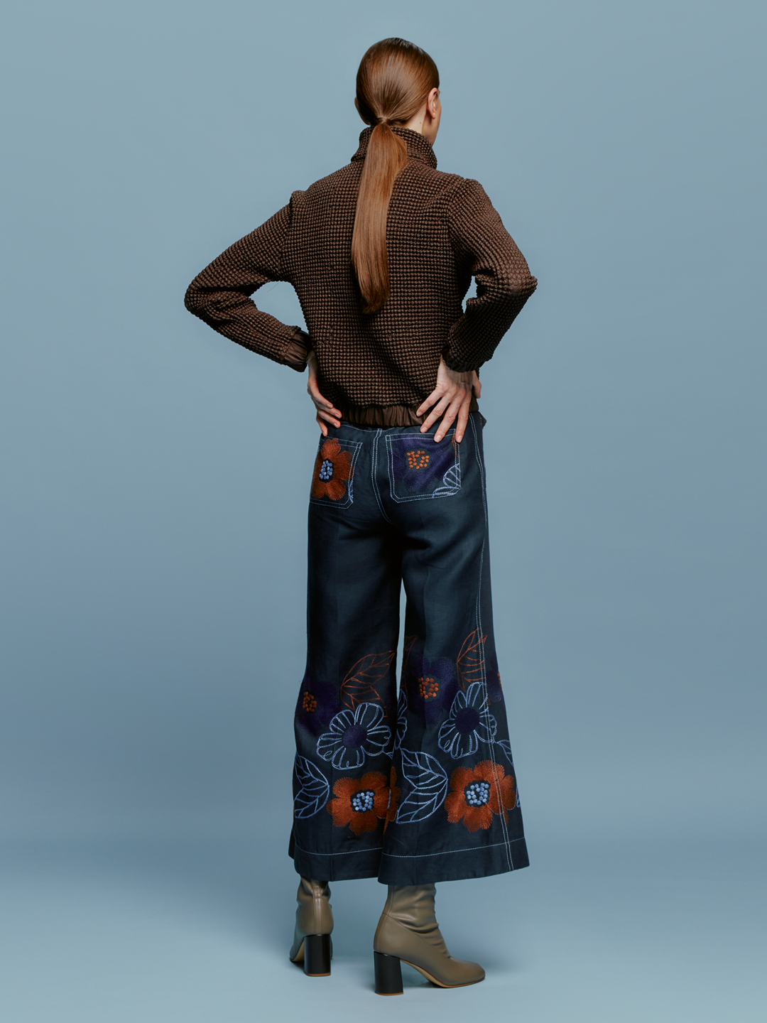 A woman stands against a plain blue background, wearing a brown sweater and the Maura Pant Navy Ochre Flower Embroidery, characterized by colorful floral patterns at the bottom. She completes her look with brown ankle boots and a neutral expression, perfectly embodying the autumn style anticipated for September 2nd, 2024.