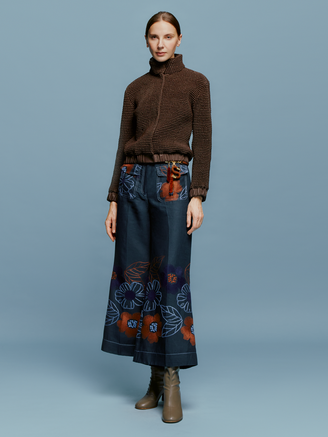 A woman stands against a plain blue background, wearing a brown sweater and the Maura Pant Navy Ochre Flower Embroidery, characterized by colorful floral patterns at the bottom. She completes her look with brown ankle boots and a neutral expression, perfectly embodying the autumn style anticipated for September 2nd, 2024.