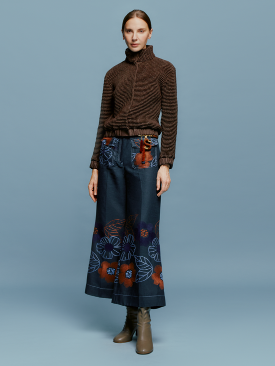 A woman stands against a plain blue background, wearing a brown sweater and the Maura Pant Navy Ochre Flower Embroidery, characterized by colorful floral patterns at the bottom. She completes her look with brown ankle boots and a neutral expression, perfectly embodying the autumn style anticipated for September 2nd, 2024.