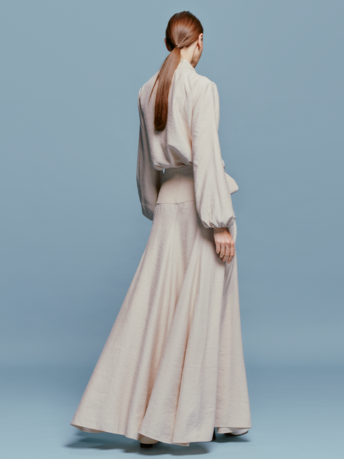 A person stands against a plain blue background, wearing a full-length, long-sleeved dress called the Modena Skirt Cream with a tie at the waist and white shoes, looking as if they are ready to embark on a ship voyage.