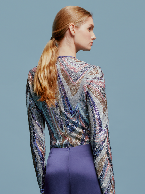 A woman with light brown hair is wearing the Nara Top Sequin Waves, a long-sleeve top featuring a multicolored wavy sequin pattern, paired with high-waisted purple pants against a plain blue background, creating an ensemble perfect for the upcoming events this August 19th.