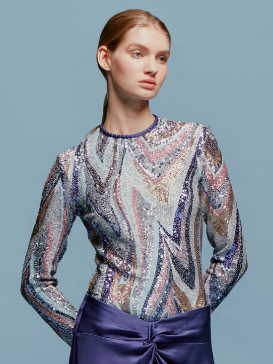 A woman with light brown hair is wearing the Nara Top Sequin Waves, a long-sleeve top featuring a multicolored wavy sequin pattern, paired with high-waisted purple pants against a plain blue background, creating an ensemble perfect for the upcoming events this August 19th.