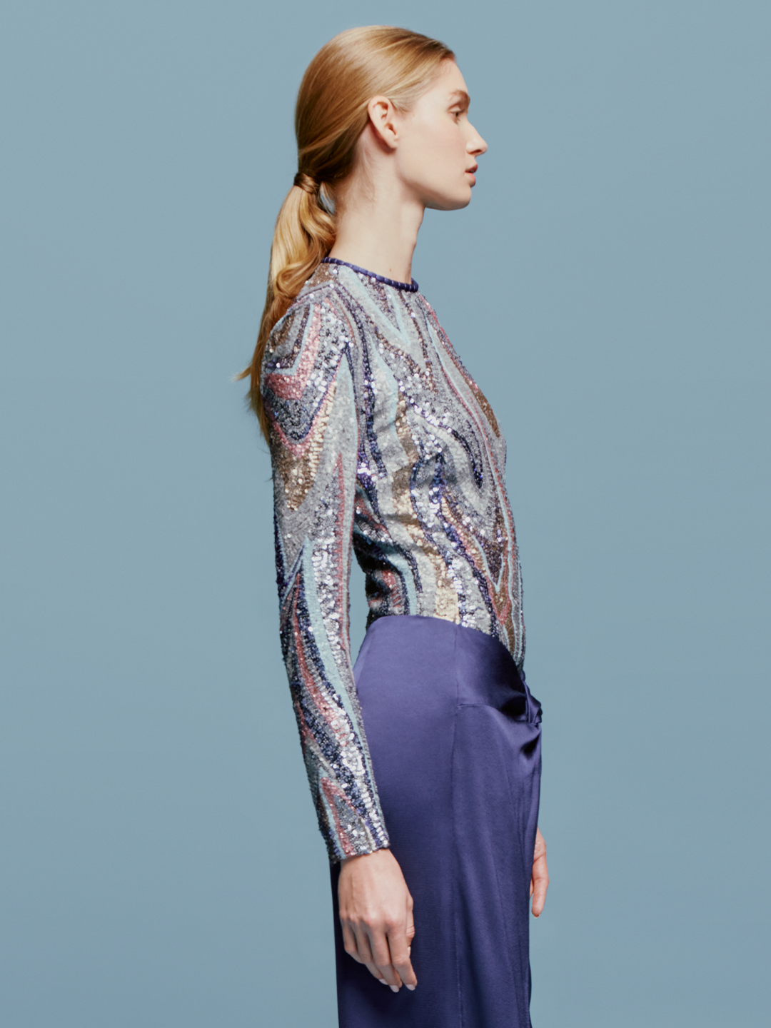 A woman with light brown hair is wearing the Nara Top Sequin Waves, a long-sleeve top featuring a multicolored wavy sequin pattern, paired with high-waisted purple pants against a plain blue background, creating an ensemble perfect for the upcoming events this August 19th.