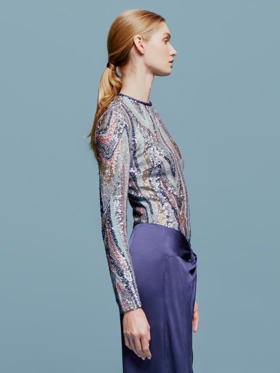 A woman with light brown hair is wearing the Nara Top Sequin Waves, a long-sleeve top featuring a multicolored wavy sequin pattern, paired with high-waisted purple pants against a plain blue background, creating an ensemble perfect for the upcoming events this August 19th.