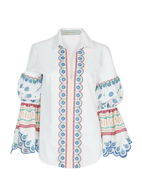 The Gaia Blouse White Blue is a white cotton piece with elaborate floral patterns and scalloped edges on the sleeves. The front features a vertical strip of intricate designs in blue, red, and turquoise, making it an exquisitely embellished garment.