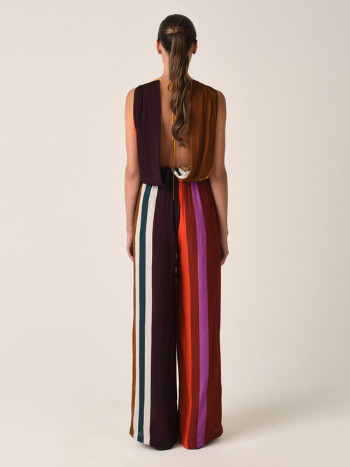 The Shelley Jumpsuit Color Block Multi is a sleeveless, halter-neck piece crafted from viscose, featuring vertical stripes in shades of mustard, maroon, red, and purple. It boasts a tie at the waist and an open back.