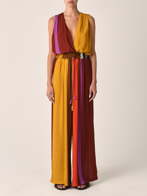 The Shelley Jumpsuit Color Block Multi is a sleeveless, halter-neck piece crafted from viscose, featuring vertical stripes in shades of mustard, maroon, red, and purple. It boasts a tie at the waist and an open back.