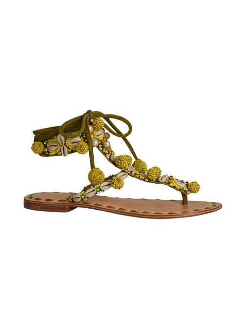 The Tisha Sandals Mustard feature a flat sandal with a brown leather sole, adorned with yellow pom-poms and beads, featuring olive green straps that tie around the ankle with added beading and seashell details.