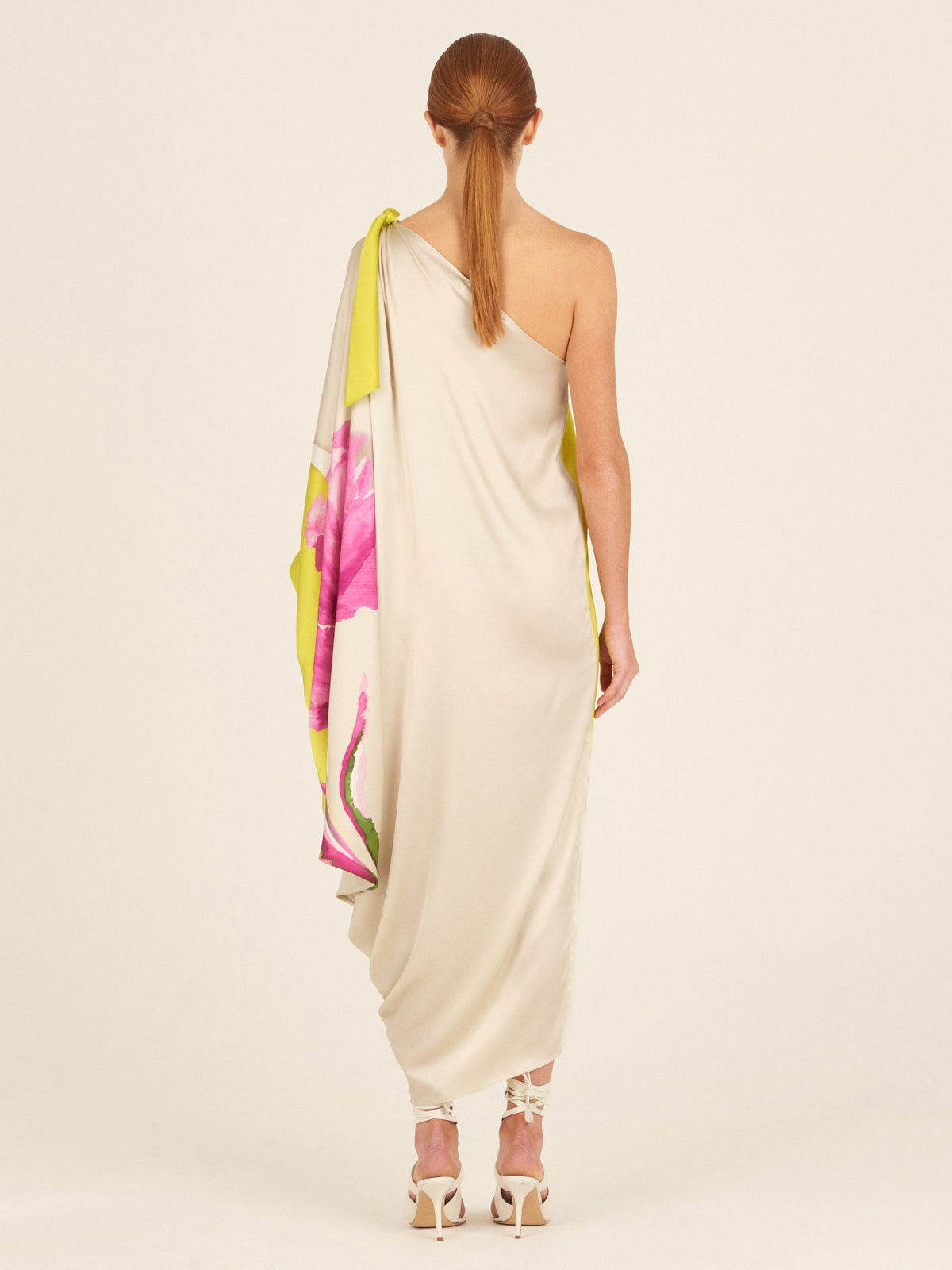 The Crema Tunic Chartreuse Beige Floral is a one-shoulder, asymmetrical silk tunic adorned with large pink floral prints and featuring a draped sleeve.