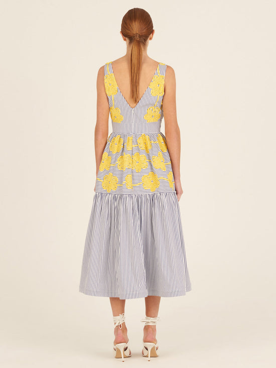 The Eugene Dress, adorned with yellow floral embroidery, showcases a sleeveless design with blue and white vertical stripes, a V-neckline, and a long-tiered skirt.